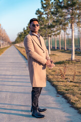 A man in a tan coat stands on a sidewalk in front of a row of trees. He is wearing sunglasses and a black belt