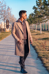 A man wearing a brown coat and black pants walks down a sidewalk. He is wearing glasses and a black shirt