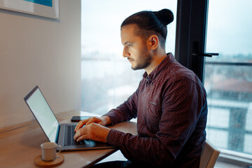 Side portrait of serious handsome man with hair bun  working at laptop, typing on keyboard,...
