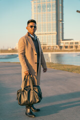 A man wearing a tan coat and holding a black bag stands in front of a tall building. The scene has a modern and sophisticated vibe, with the man dressed in a stylish outfit © oybekostanov