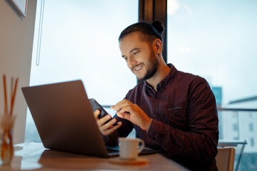 Portrait of smiling handsome man working at laptop while using smartphone on background of panoramic window.