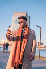 A man is taking a selfie with his cell phone while wearing a brown coat and an orange scarf