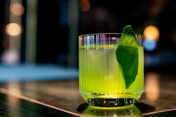 A vibrant green cocktail with a fresh leaf garnish, served on a bar counter with blurred lights in...