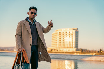A man wearing a brown coat and sunglasses is standing on a sidewalk next to a body of water. He is...