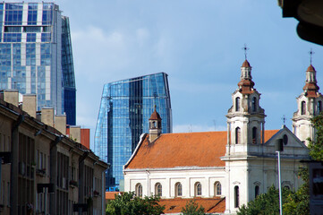 Obraz premium An old religious building, a cathedral with bright orange roof tiles, towers, columns, against which the blue sky and tall buildings belonging to modern architecture with glass facades