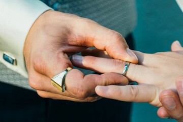 Person's hand gently placing a shining gold wedding ring on the ring finger of another person