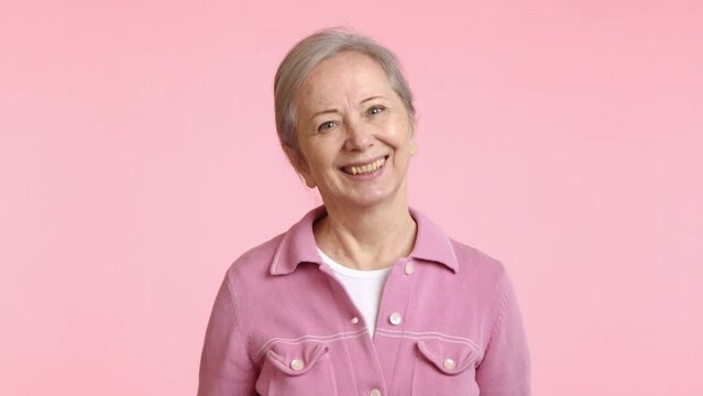 A senior woman with a knowing look makes the 'zip your lip' gesture, her hand motion signifying secrecy or the need for silence, in front of a gentle pink background. Camera 8K RAW.