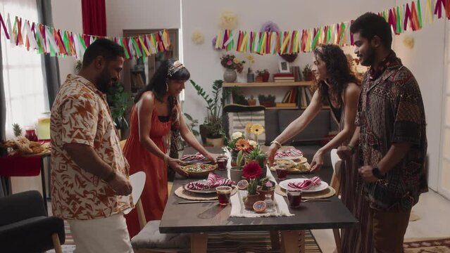 Full zoom footage of four young Hispanic men and women in bright colorful shirts and dresses bringing platters with traditional Mexican foods and cocktails to table for sumptuous holiday feast