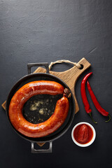 Ring of fried pork sausage on a black cast iron frying pan with hot pepper and ketchup on a black background. Top view with copy space