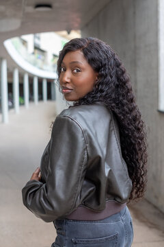 In an urban underpass, a young African woman with long curly hair turns to glance over her shoulder. She exudes casual confidence and style in a sleek leather jacket, against a backdrop of concrete