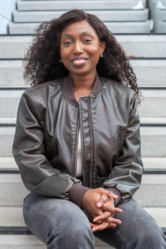 A young African woman with a radiant smile sits comfortably on outdoor steps, her dark curly hair framing her face. She wears a fashionable leather jacket, presenting a relaxed yet confident posture