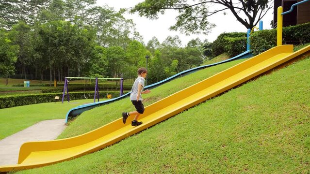 With boundless energy, boy joyfully climbs up plastic slide chute, showcasing his agility and strength. Young boy dashes around playground, his movements captured in slow motion from side view. Active