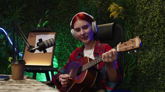 Gen Z girl in headphones playing guitar and singing in microphone while recording music in modern studio with neon illumination and green walls