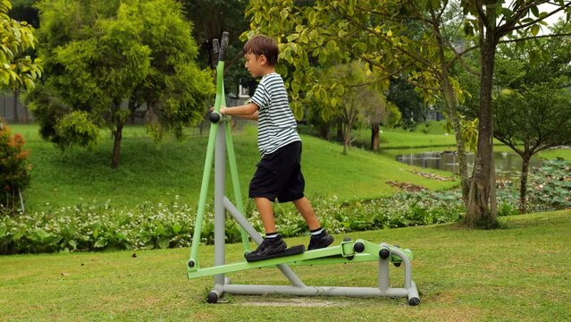 In green park, boy exercises on single ski stepper machine, playfully discovering his motion. Standing tall, he holds onto horizontal bar, stepping with youthful energy. This outdoor gym workout