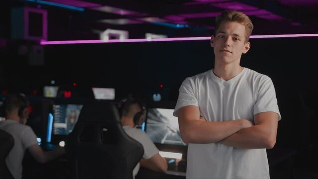 Portrait smiling Caucasian youth gamer teenager poses, player crosses his arms and looks at camera in computer club, cyber sport tournament on PC. E-sport multiplayer online video games championship
