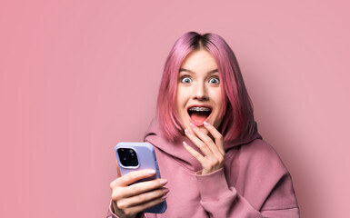 Shocked astonished very happy pink hair woman wear braces, hoodie opened mouth, using holding hand cell phone, modern smartphone, mobile phone, isolated wall background. Online offer, dental care ad. - obrazy, fototapety, plakaty