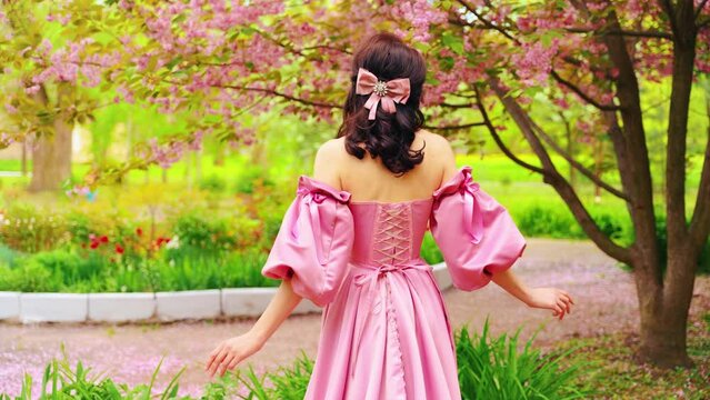 Fantasy noble Woman queen in spring garden pink flowers tree brightg reen grass summer park. girl in long pink dress medieval lady vintage old style bow ribbon in hair back rear view enjoy nature 4k