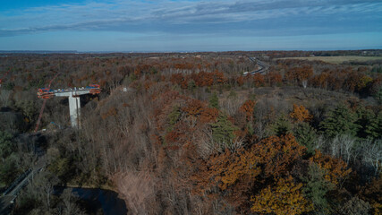 An aerial view of a bridge being built over a valley with a river and Forest in the fall