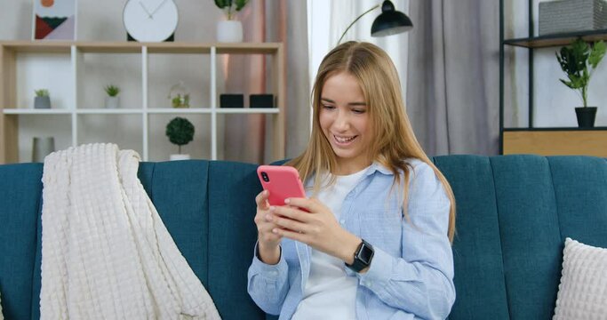 Blondie woman in casual clothes writing message on phone to her friend sitting on soft sofa in living-room