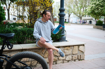 Athletic young man cyclist relaxing when riding electric bike, holding a bottle of water while...