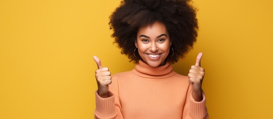 A woman with a big afro hairstyle is smiling with her nose, eyebrows raised, giving two thumbs up gesture, showing happiness on a yellow background