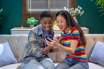 Black and asian friends are sharing a moment of leisure on a couch by a window, smiling at a cell phone excited and surprised. They look happy and comfortable, with a houseplant in the corner of the