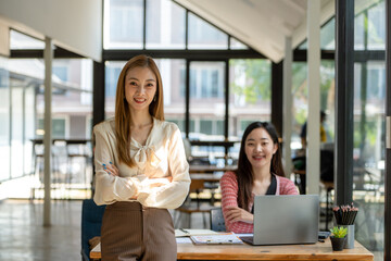 Obraz premium A confident businesswoman stands arms crossed in a modern collaborative workspace with a seated colleague in the background.