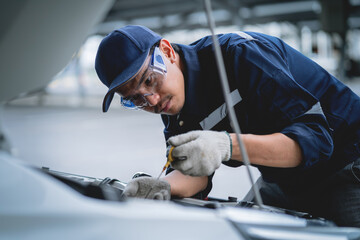 A mechanic is working on a car engine, wearing a blue shirt and a blue hat. He is wearing gloves and goggles, and has a wrench in his hand