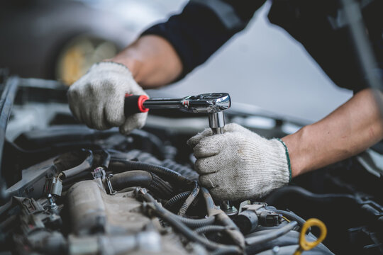 A man is working on a car engine, wearing a blue hat and safety glasses. He is using a wrench to loosen a bolt