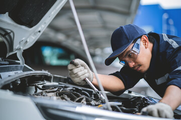 A man is working on a car engine, wearing a blue hat and safety glasses. He is using a wrench to...