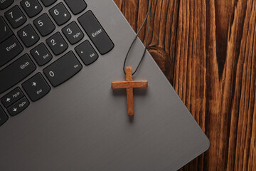 Wooden Christian cross on a string with laptop, wooden table. Online Christian Education