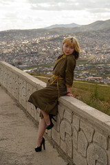 Fashionable young woman with blonde hair wearing a patterned dress and high heels, posing elegantly on a stone ledge with a panoramic view of a cityscape in the background at dusk.