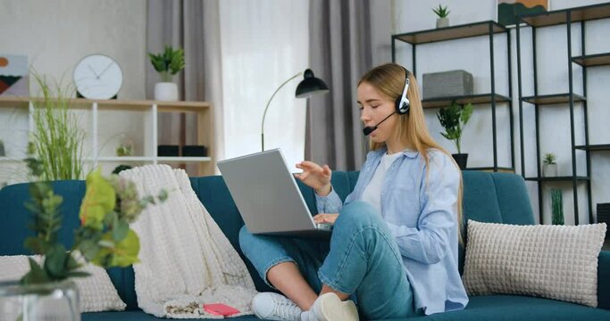 Likable woman in headset has online video conference with friend sitting in lotus pose on couch at home