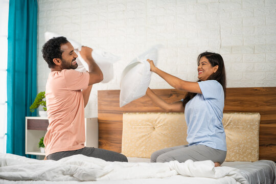 Playful indian couples fighting with pillows at bedroom - concept of intimacy, flirting and joyful relationship bonding