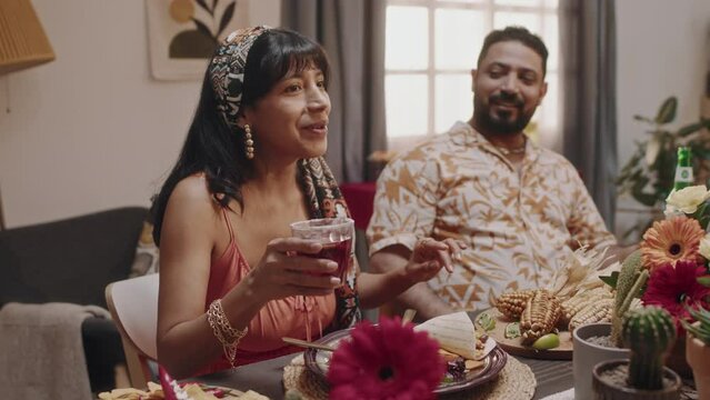Medium shot of beautiful young Mexican woman with bright scarf on hair, in strappy dress sitting at party table, holding cocktail, telling funny story to husband and friends, laughing