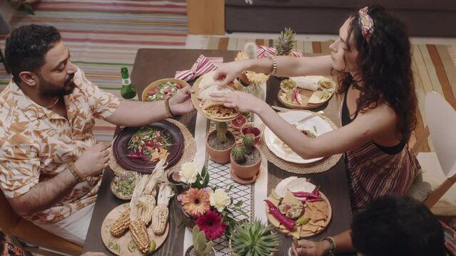 Medium full top shot of two young Mexican couples sitting around festive table while celebrating Cinco de Mayo, eating tasty traditional foods, husband passing over plate of chalupas, wife taking one