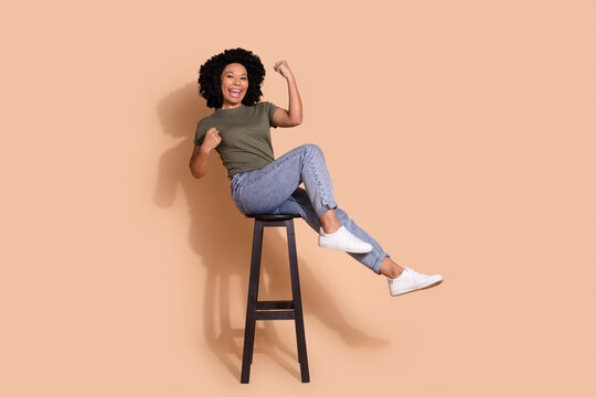 Full length photo of overjoyed girl sit on chair clenching fists win lottery shout win gambling isolated on beige color background
