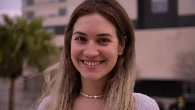 Close up of cheerful blonde girl outdoors. European woman poses looking at camera smiling and nodding her head. Happy people posing for video with city buildings in background.