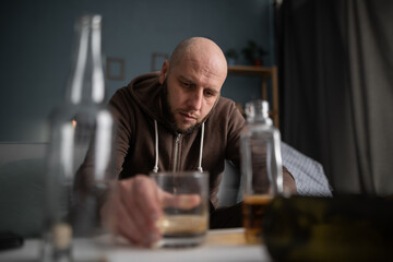 Drunk man or male alcoholic drinking whiskey at home sitting between many bottles. Alcoholism,...