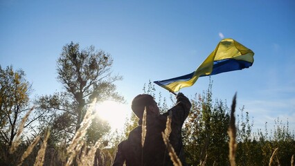 Male military in uniform waving flag of Ukraine at countryside. Young soldier of ukrainian army lifting blue-yellow banner as symbol of victory against russian aggression. Invasion resistance concept.