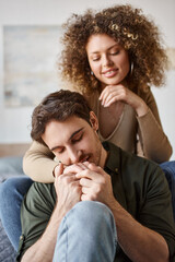 Curly woman hugging her relaxed boyfriend from behind sitting on bed smiling together