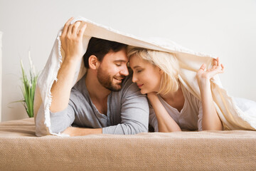 Portrait image - young inlove couple with closed eyes, covered with sheet-blanket over heads, lying laying on bed. Blond woman, brunette bearded man at home. Love, relationship, happy family, dating.