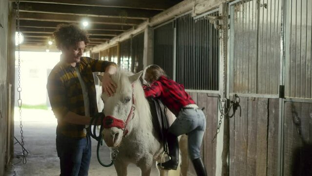 Little girl child climbs up on to a horse in a barn on the family ranch