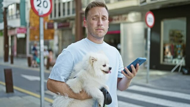 Cheerful young caucasian man enjoys bond of happiness outside, confident, texting on smartphone while smiling wide, standing with pet dog on the lively city street.