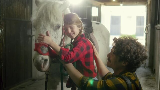 A little girl strokes and kisses her pet horse while spending time in the barn with her farmer father