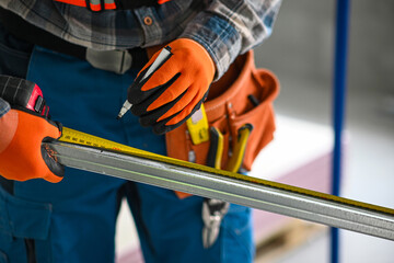 a construction worker in overalls, gloves and a tool belt measures the length of a plasterboard...
