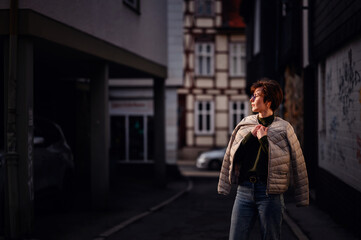 A fashion-forward woman with glasses and a green sweater carries her jacket confidently in a dimly lit urban alley.