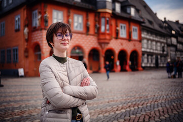 A woman stands confidently in a historic town square, her arms crossed, with traditional...