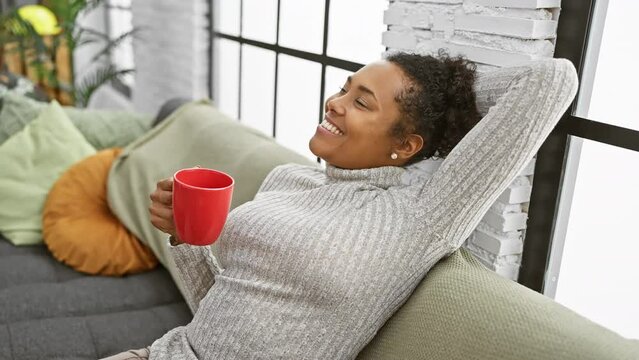 Relaxed woman with curly hair smiling and holding a red mug in a cozy living room
