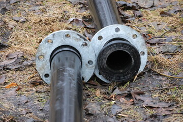 Unconnected black heavy-duty plastic pipes for water pumping with joints constructed of metal. Clear bolts and screws. The pipes are located on the ground in nature.
